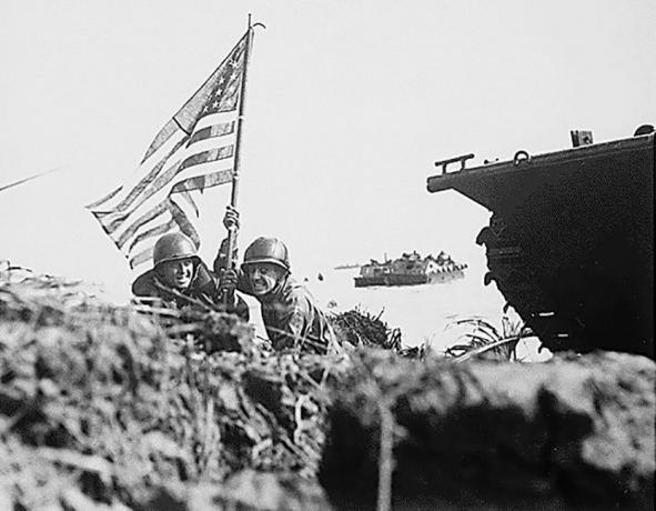 To soldater med et amerikansk flag på en strand ved siden af ​​et sporet køretøj.